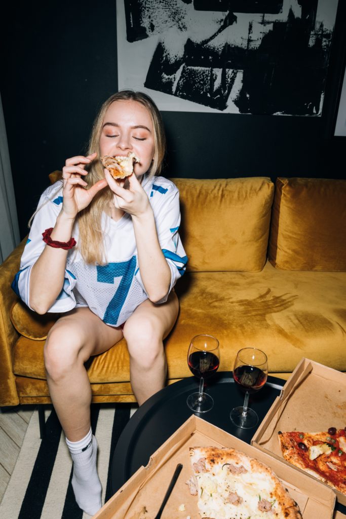 Girl eating pizza and drinking wine while sitting on a couch - christmas party ideas
