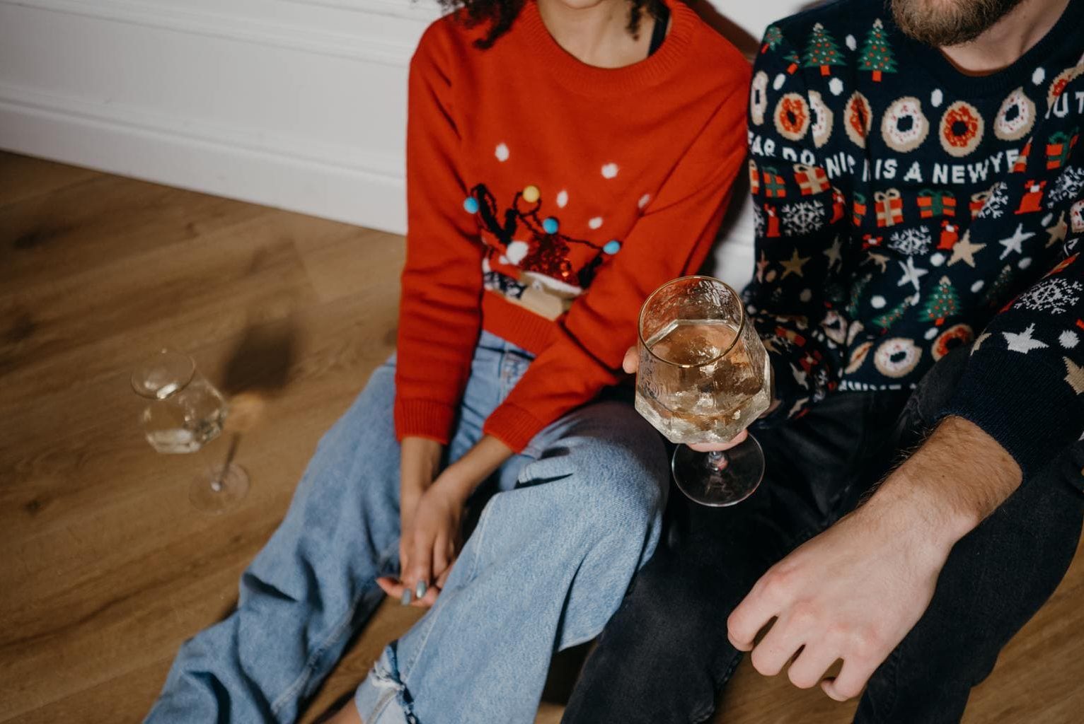 Two people wearing holiday sweaters sitting on the floor and drinking wine.