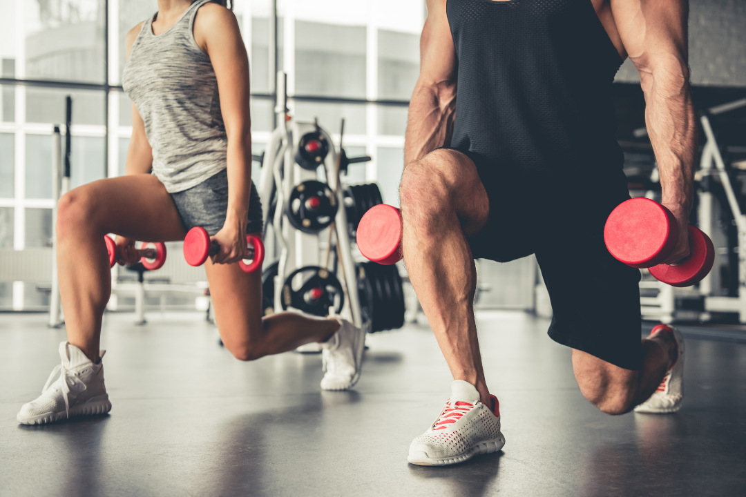 Photo of a man and woman at a gym doing lunges while holding dumbbells