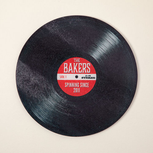 Black glass cutting board in the shape of a record with a red label and text The Bakers, Spinning Since 2011 in the center