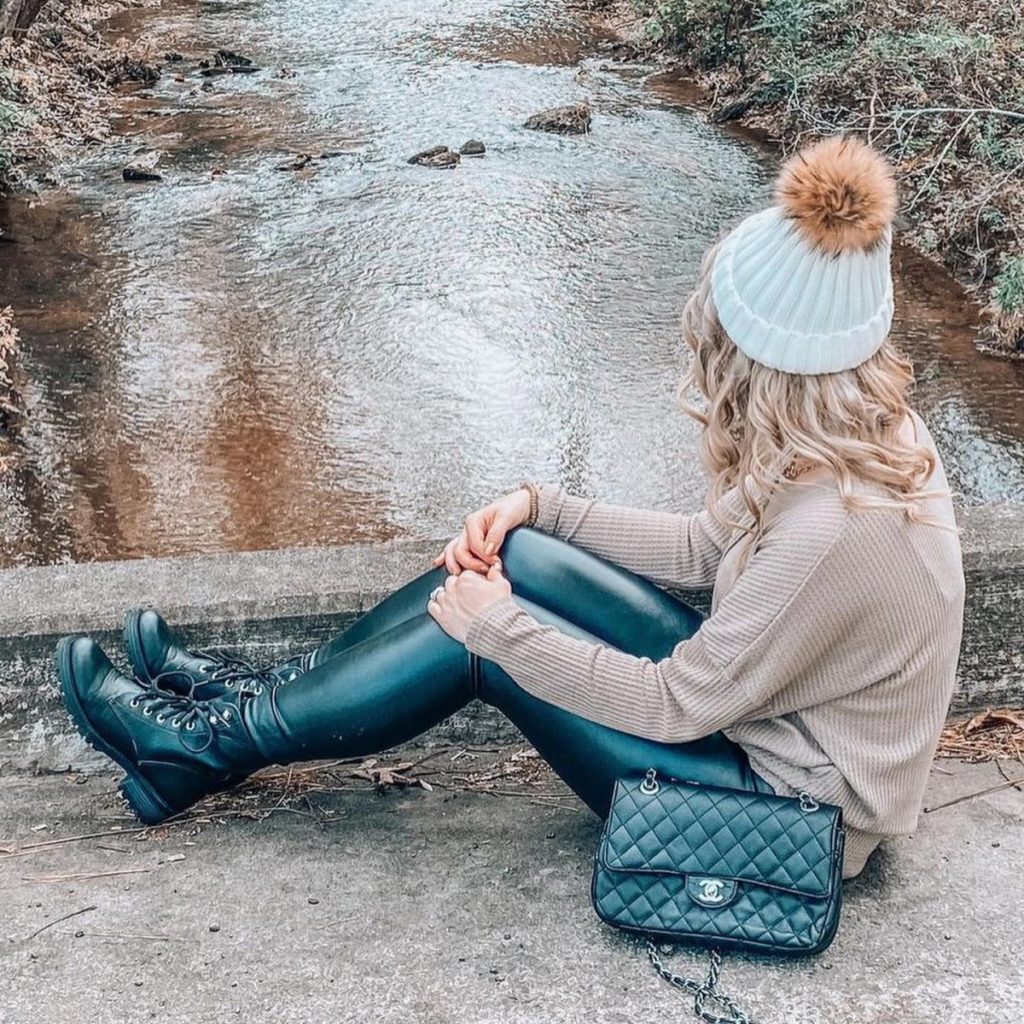 Photo of a woman sitting by a river wearing leather pants, combat boots, a chain strap bag, and a sweater