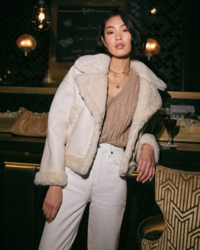 White jeans outfit for winter with white furry moto jacket, light brown sweater, layered necklaces, ankle boots