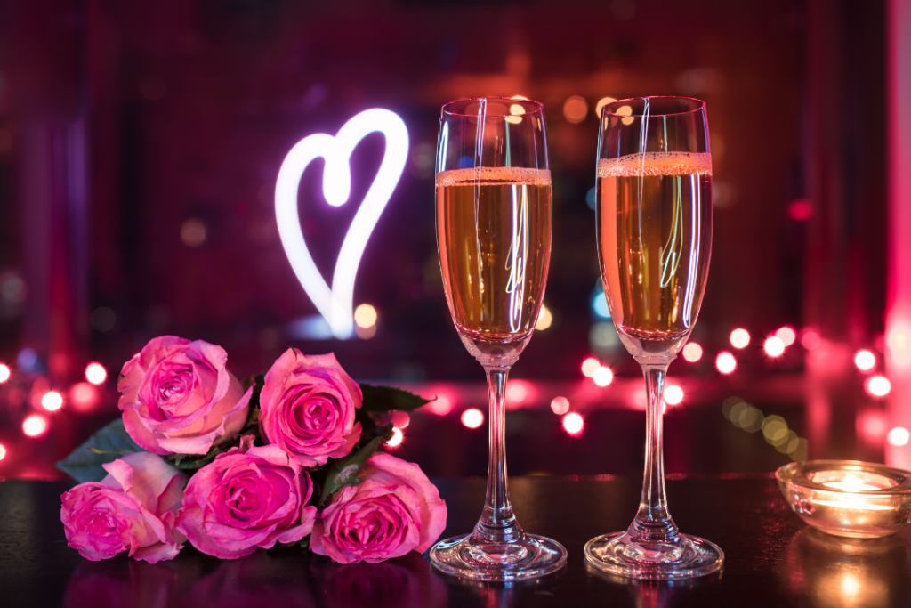 Photo of two glasses of champagne next to some roses with a neon heart sign in the background