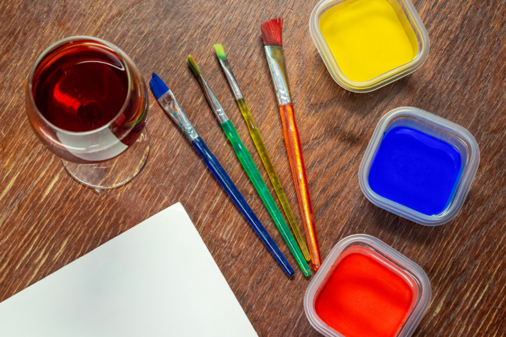 Photo of paints and paintbrushes laid out next to a glass of red wine
