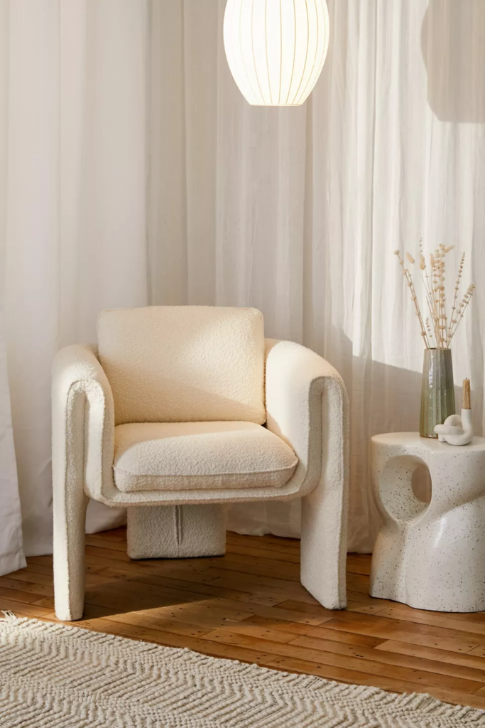 Textured cream velvet chair in front of white curtains, next to a cream rug and a white speckled side table