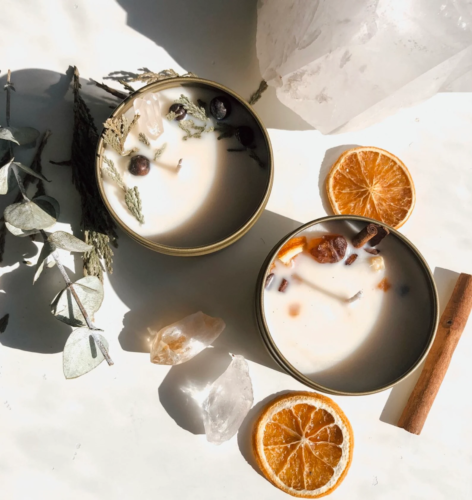 Photo of a DIY candle making kit with two candles surrounded by cinnamon sticks, herbs, eucalyptus, dried oranges, and crystals