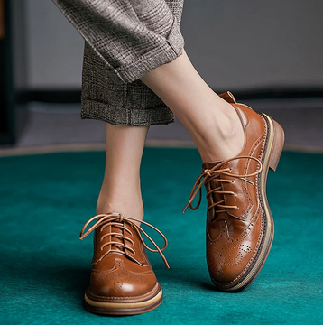 picure of model from the calves down with her feet crossed wearing neutral pants and western looking oxfords that have no heel on green teal background