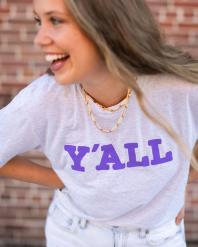 Girl wearing a Mardi Gras t-shirt that says Y'all, along with light was jeans and gold jewelry