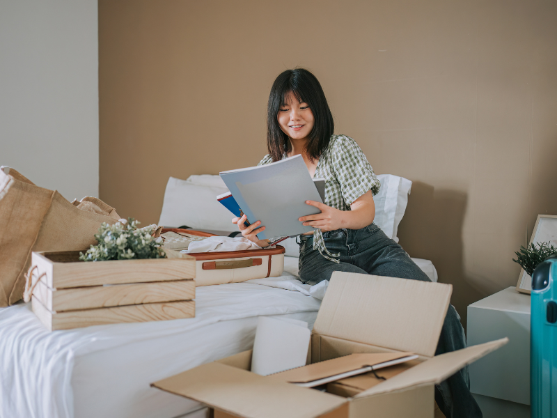 College student sitting on her bed in her apartment, unpacking her decor