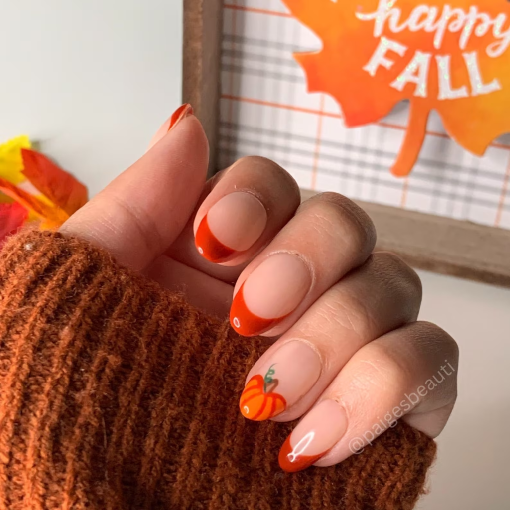 Photo of a woman in a burnt orange sweater modeling a fall manicure consisting of nude nails with orange french tips and a pumpkin accent nail on the ring finger