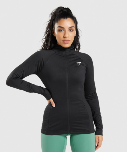 Photo of a dark haired woman wearing a Gymshark black zip-up training jacket with green leggings on the bottom
