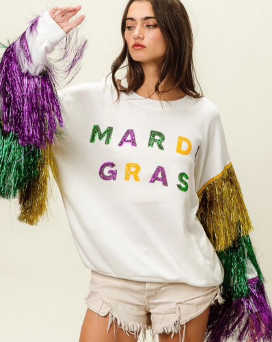 Woman wearing a white sweatshirt that reads Mardi Gras in glitter letters, with sleeves decorated in green, purple, and gold fringe. The sweatshirt is worn with beige distressed denim shorts.
