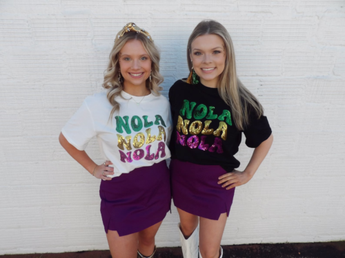 Photo of two girls wearing t-shirts that read NOLA NOLA NOLA in sequin mardi gras colors. One girl wears a white version of the shirt and the other wears a black. Both pair their tees with purple mini skirts and boots.
